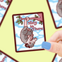 Hand holding a Hang In There Opossum Sticker featuring a cute possum hanging from a tree branch with blue sky background.