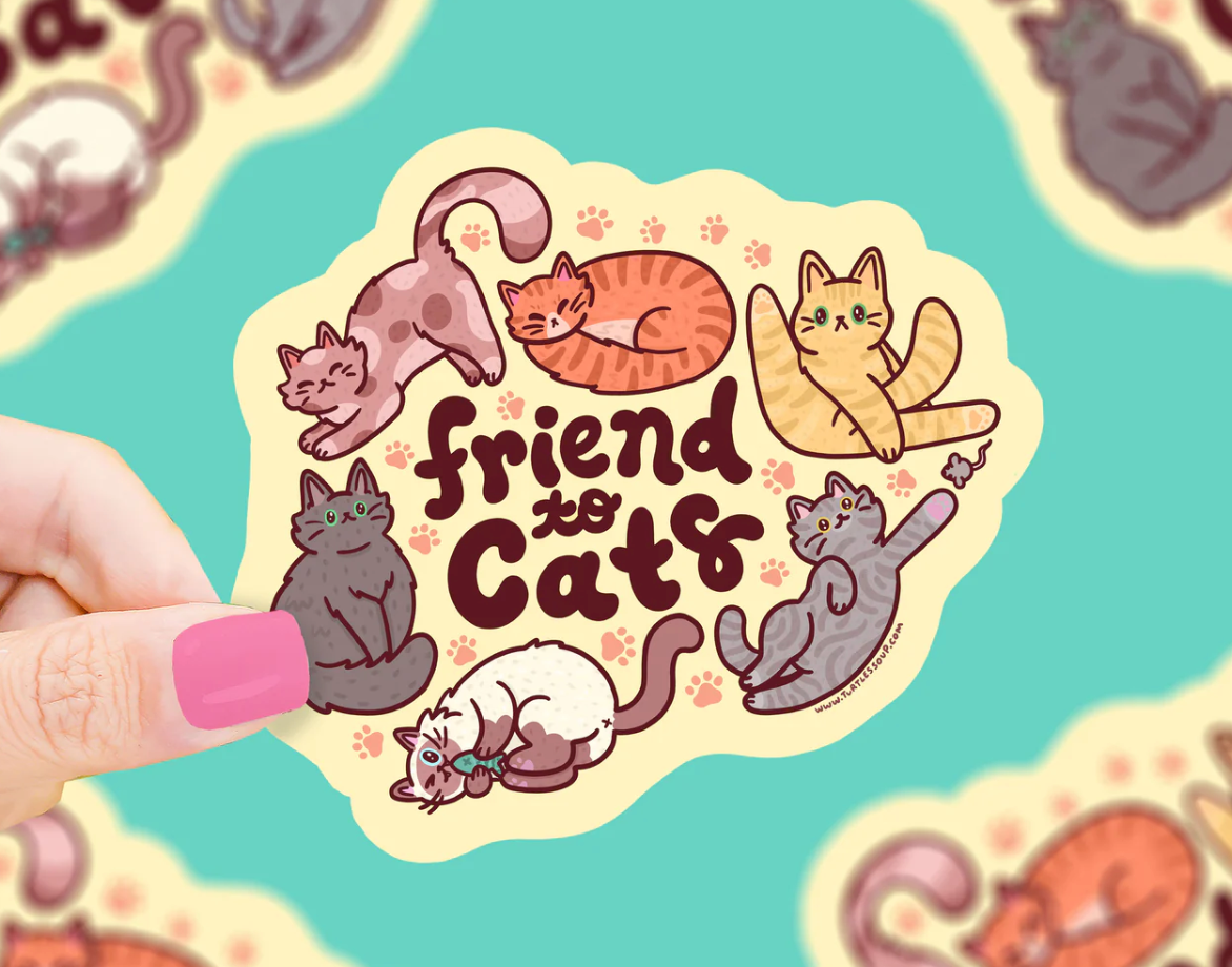 Hand holding a colorful Friend To Cats Sticker featuring six playful cats and paw prints on a cream background.