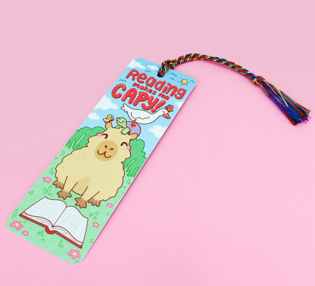 Bookmark | Reading Makes Me Capy with cute capybara reading book and rainbow tassel on pink background.