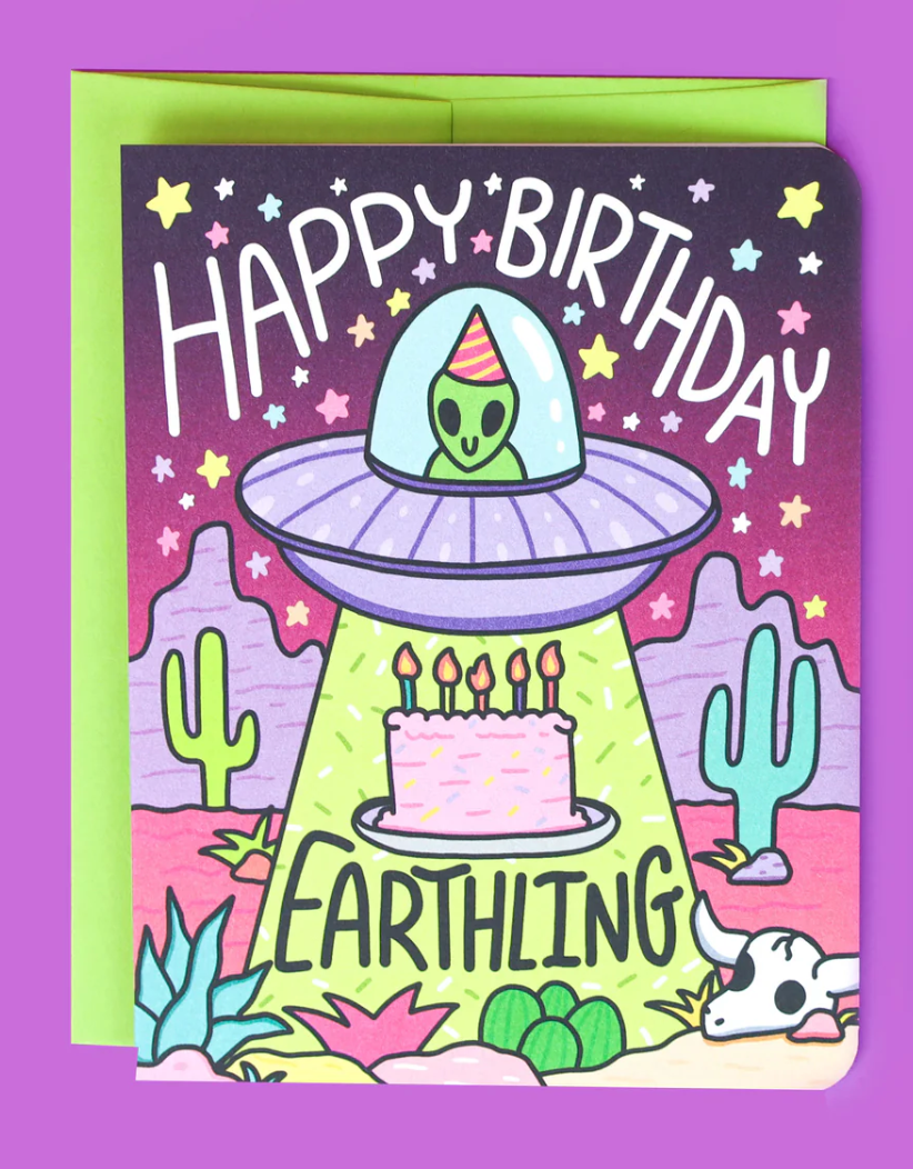 Happy Birthday Earthling Card with alien in UFO delivering birthday cake on colorful desert background.