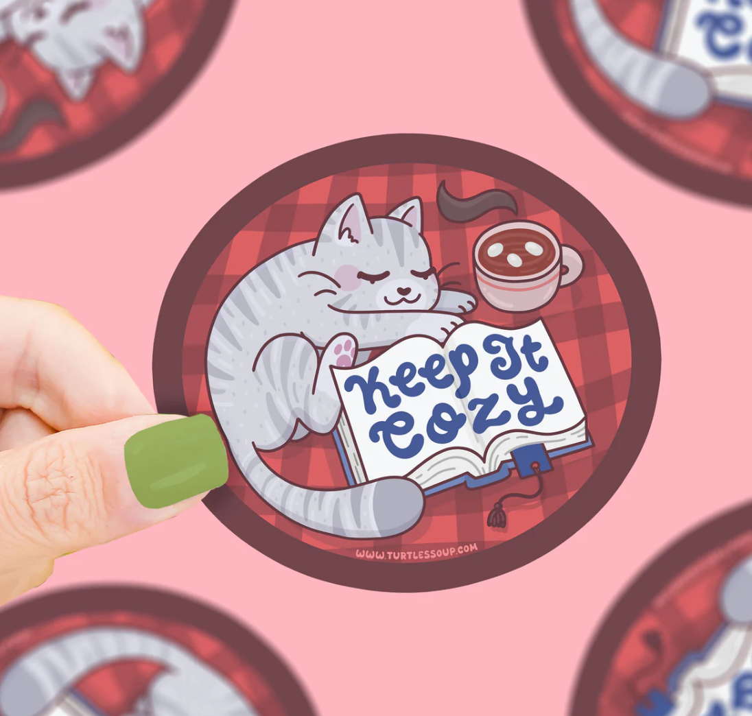 Hand holding Keep It Cozy Sticker with a sleeping gray cat, open book, and cup of cocoa on a red plaid background.