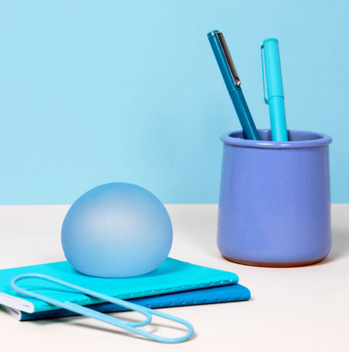 Speks. Gump Dew stress ball on a notebook beside a pen holder with two blue pens on a desk.