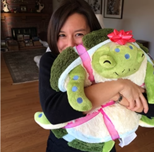 Sea Turtle plush toy hugged by smiling woman, featuring green shell, blue spots, and a red bow on its head.