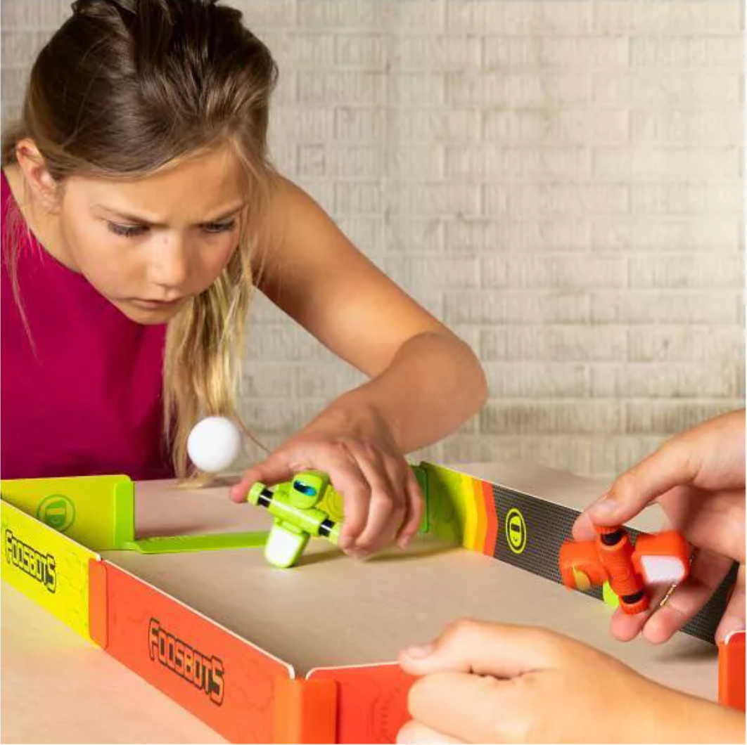 Girl playing with Foosbots Stadium Battle Set, controlling green and orange Foosbots in a tabletop soccer game.