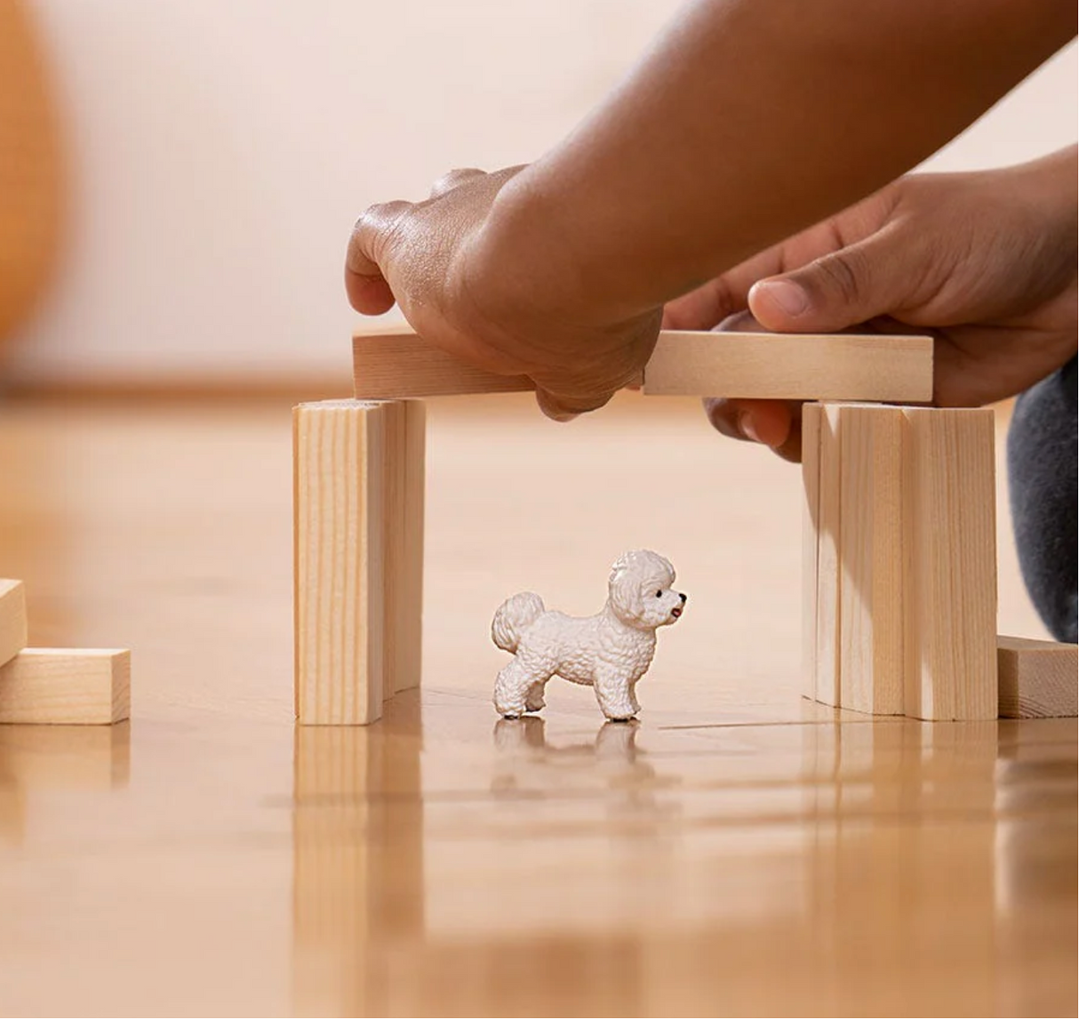 Schleich® Bichon Frise figurine stands on wooden floor surrounded by hands building a wooden block structure.