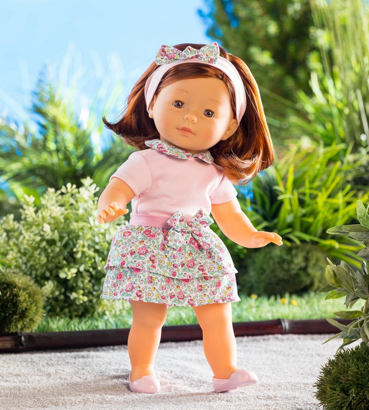 Ma Corolle Pia doll with silky red hair, pink t-shirt, floral skirt, headband, and ballet flats in garden setting.
