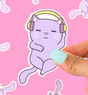 Hand holding a purple cat sticker wearing headphones, titled Headphones Cat Sticker, against a pink background.
