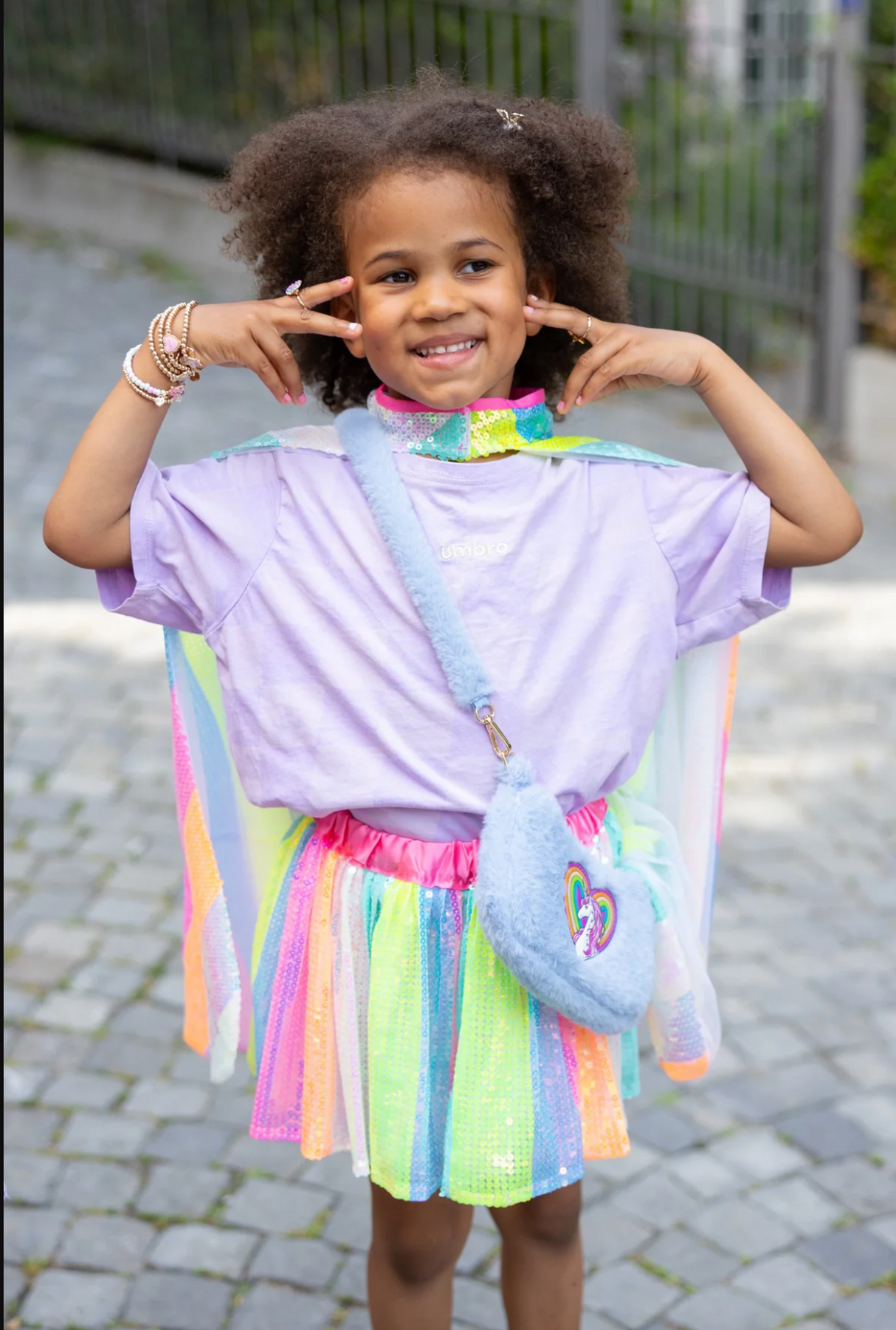 Rainbow Cuddle Purse | Periwinkle worn by a smiling child in colorful outfit with unicorn and rainbow heart design.