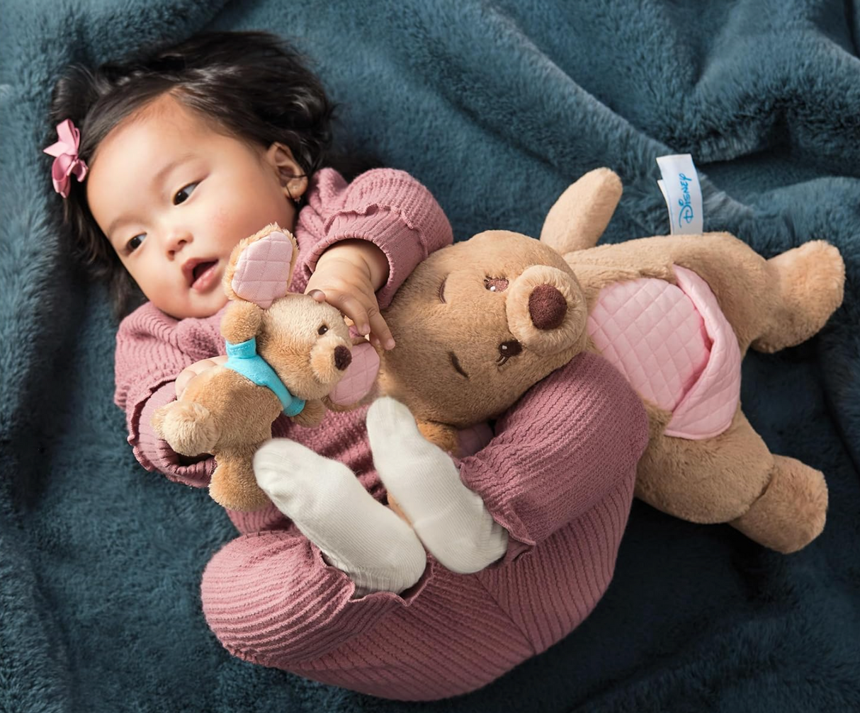 Baby lying on blue blanket cuddling Oh So Snuggly Kanga & Roo plush toys with soft quilted accents and embroidered details.