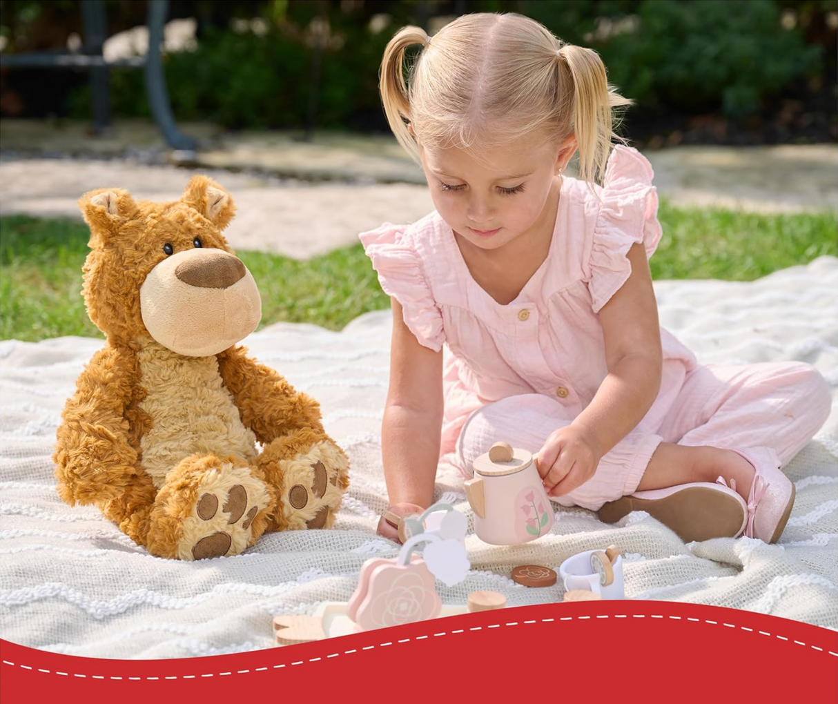 Teddy Bear Bexley seated on blanket next to a toddler playing with wooden tea set outdoors on a sunny day.