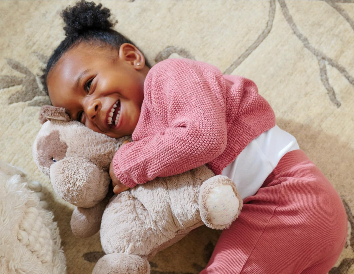 Child lying on carpet smiling and hugging an Oh So Snuggly Cow plush toy with mocha spots and soft quilted details.