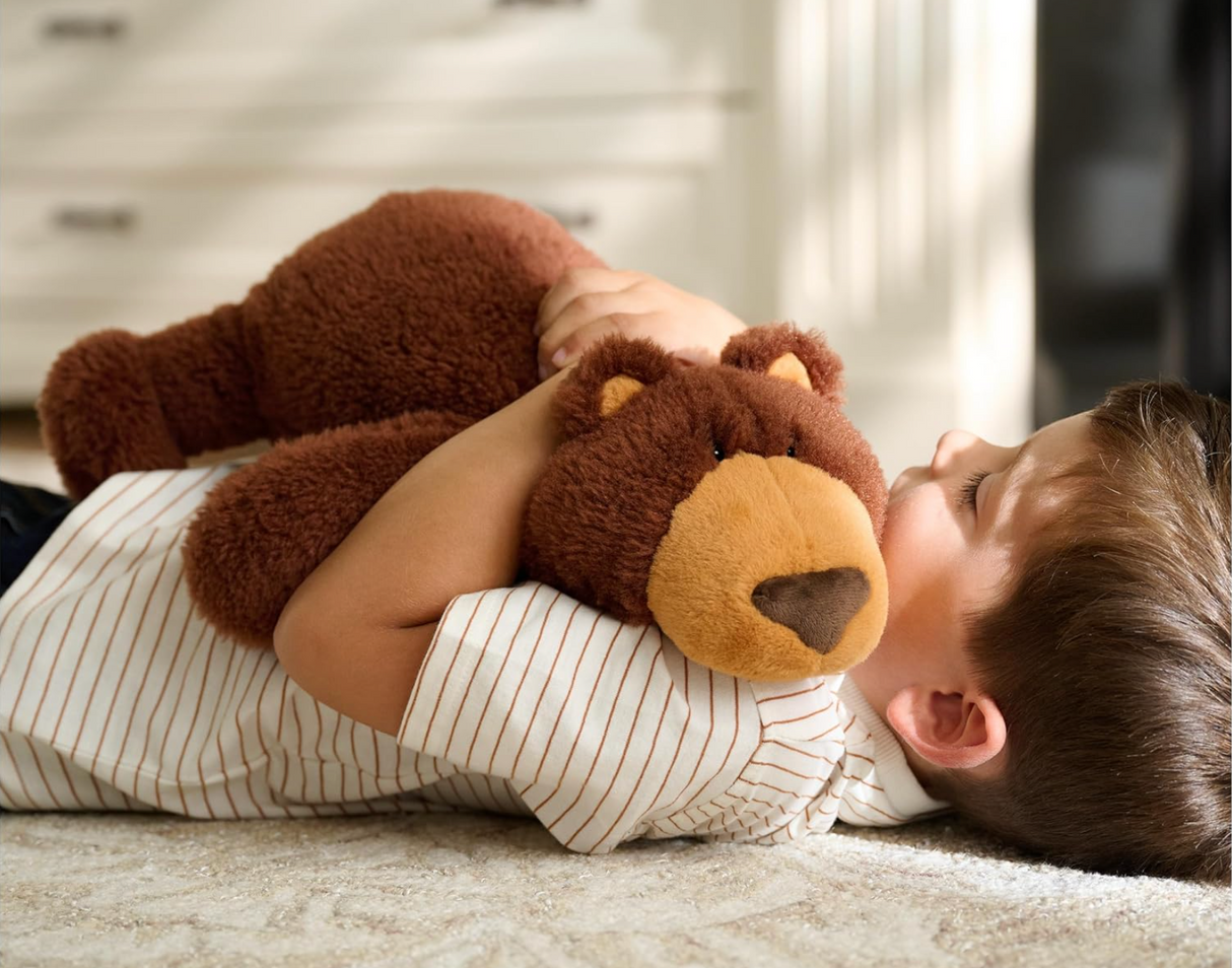 Child lying on floor hugging a fluffy chestnut-brown bear in a relaxed pose, Teddy Bear Tush plush toy.