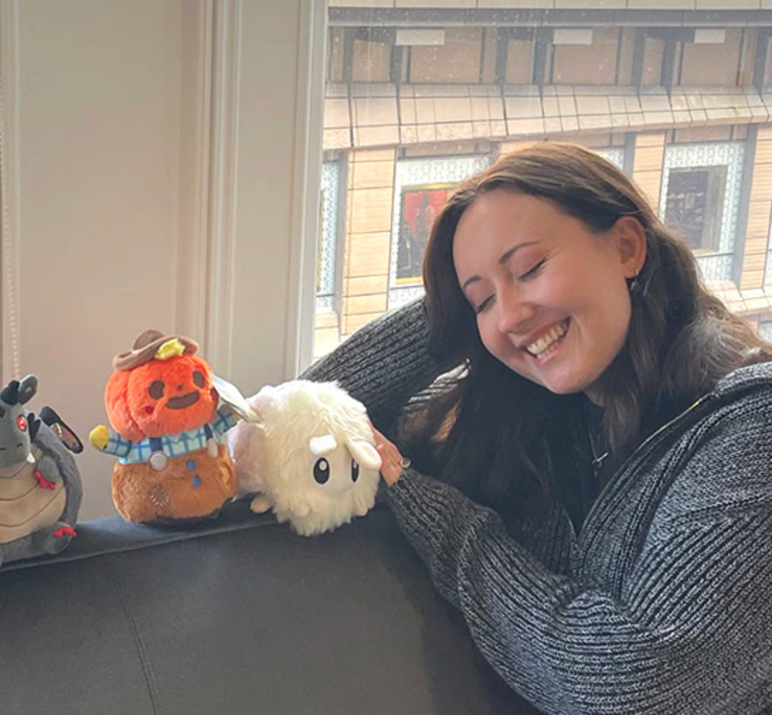 A smiling woman interacts with Poodle Moth Snackers plush toy on a couch by the window.