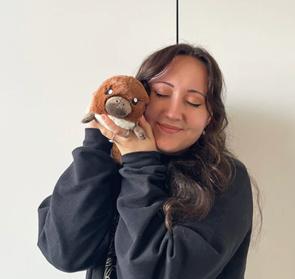 Woman smiling while holding a soft Platypus Snackers plush toy close to her face against a plain background.