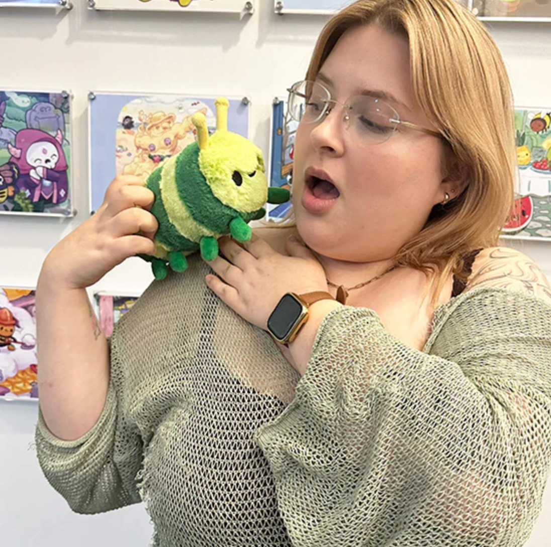 Woman holding a green and yellow plush toy smiling surprised with Caterpillar Snackers on her shoulder.