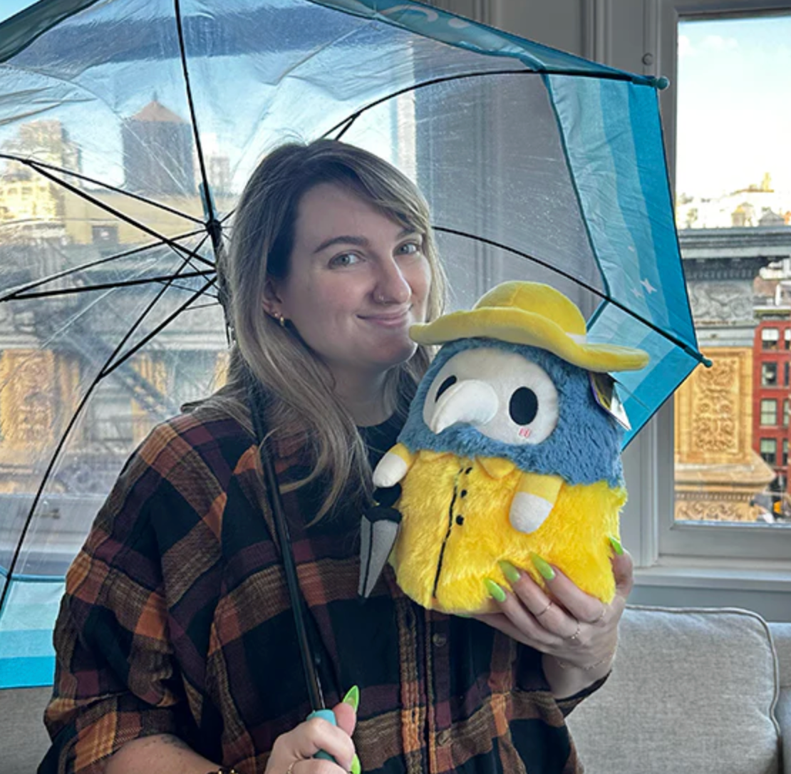 Woman holding Alter Ego | Plague Doctor Rainy plush toy under a blue umbrella by a window with city view.