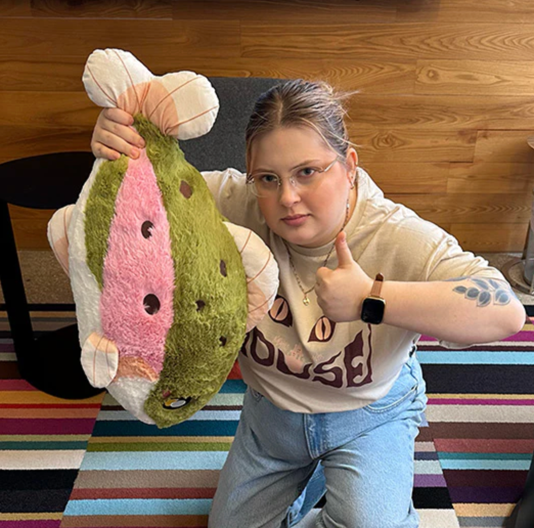 Person holding a large Rainbow Trout plush giving a thumbs up, sitting on a colorful striped rug.