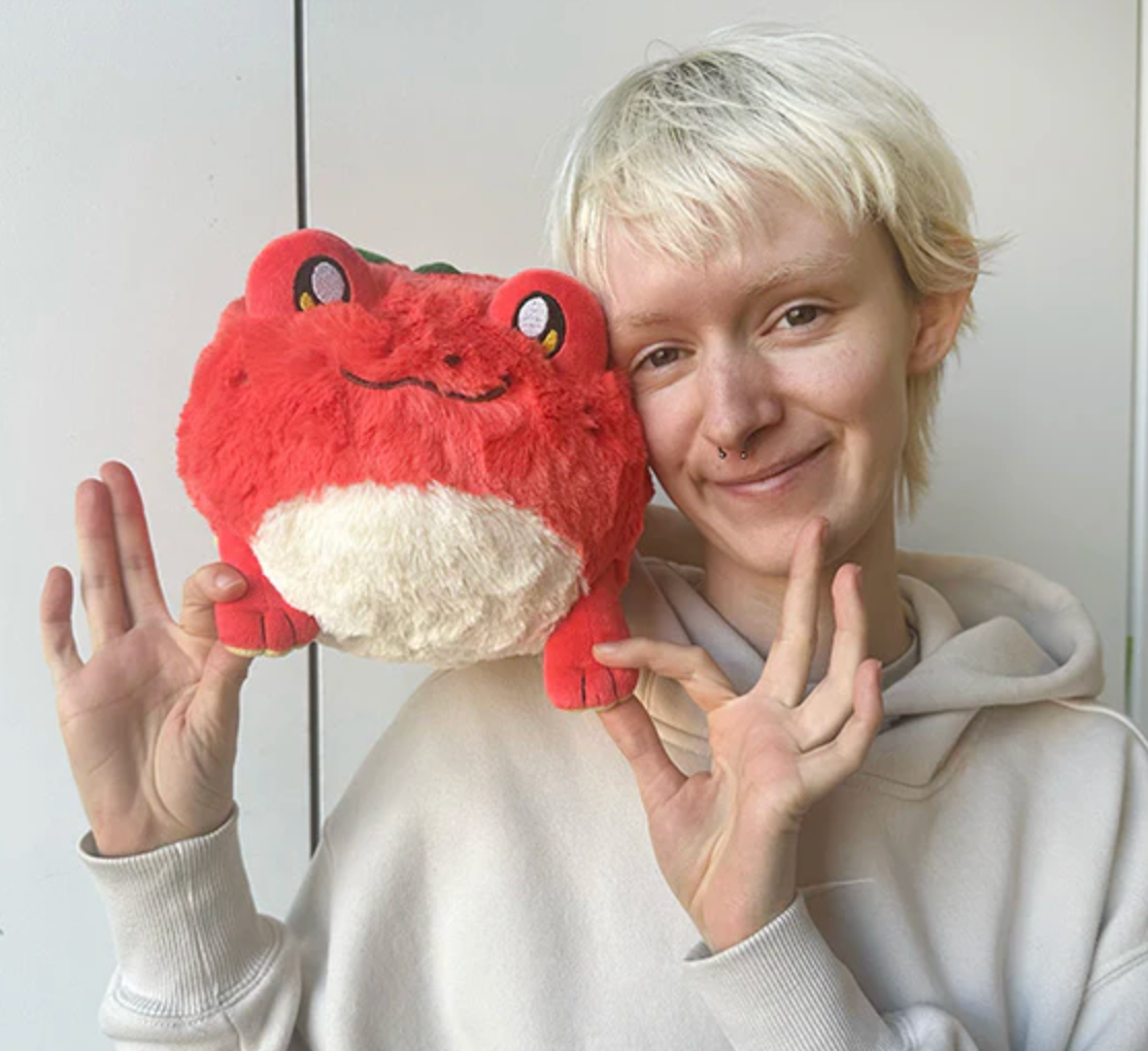 Person holding a bright red and white plush TomAToad Mini with a friendly smiling face.