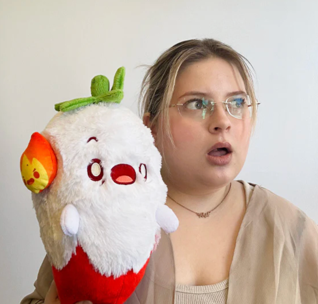 Woman holding a surprised expression while holding a Ghost Pepper Mini plush toy with fiery headphones.