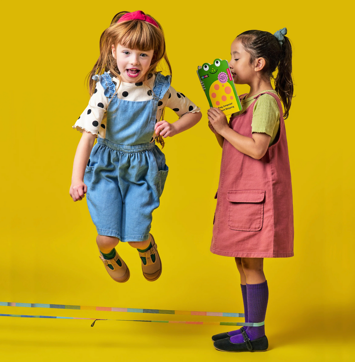 Two children playing with Frog Jumpsies! French Skipping rope and booklet on bright yellow background.