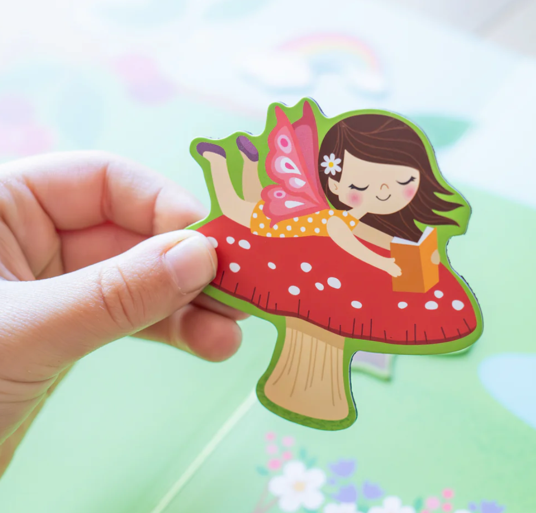 Hand holding a fairy magnet piece from the Magna Carry Forest Fairies set showing a girl reading on a red mushroom.
