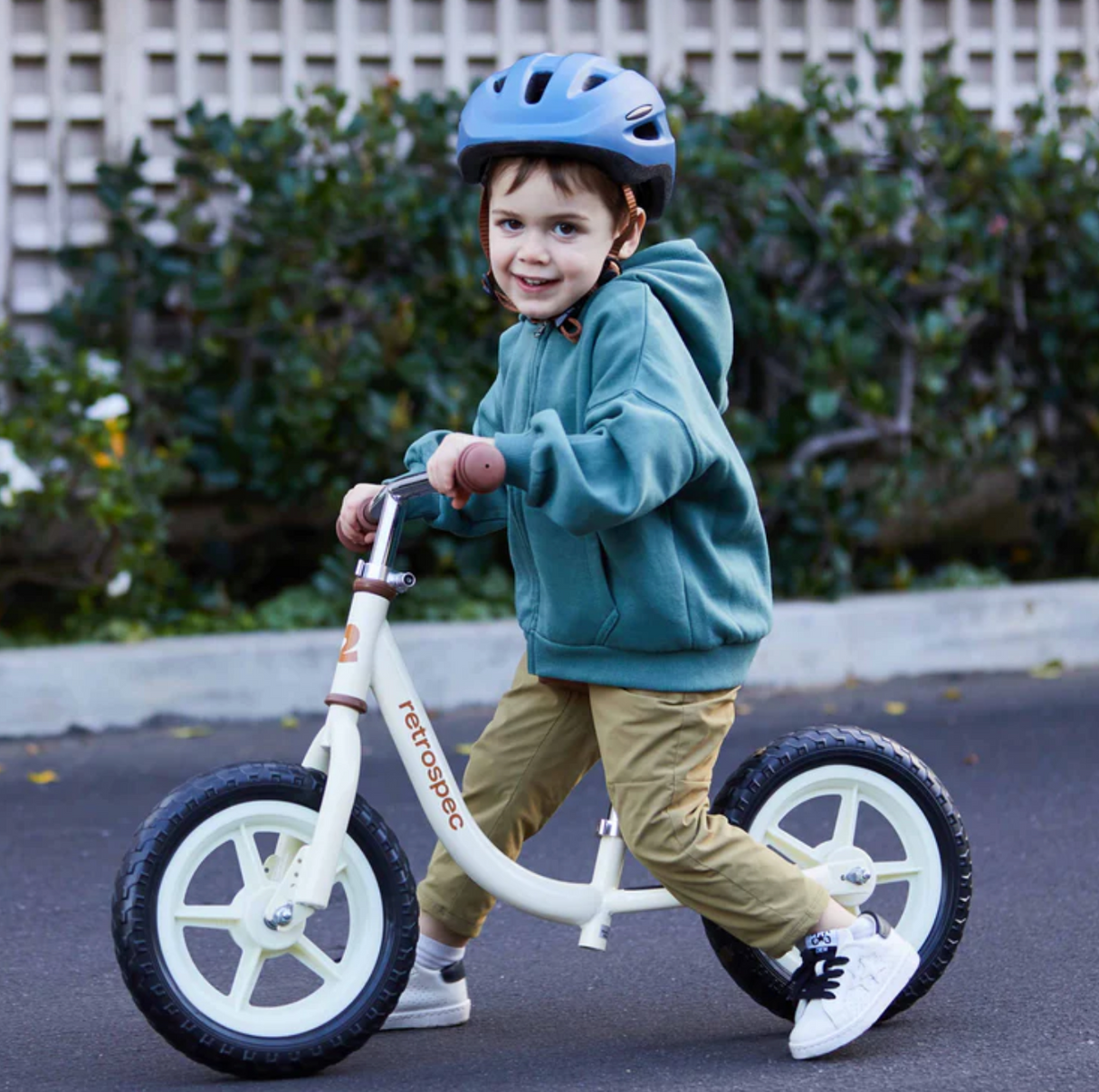 Child wearing a helmet riding the Cub Balance Bike | Blush outdoors on a paved path.