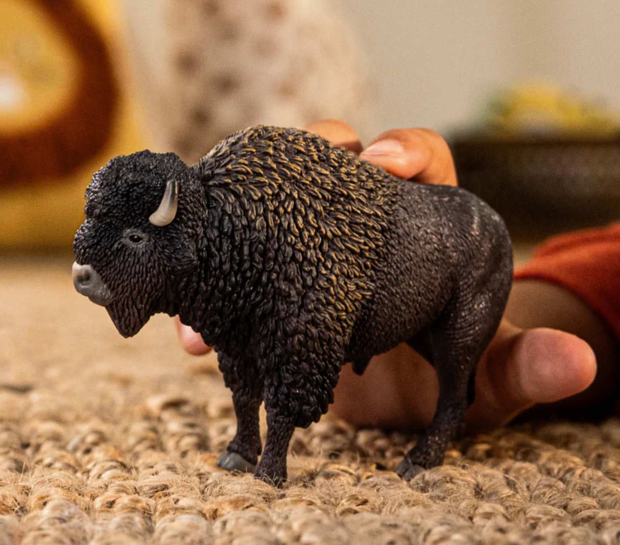 Bison toy with a mighty head, distinctive hump, and thick brown coat, held by a child's hand on a textured surface.