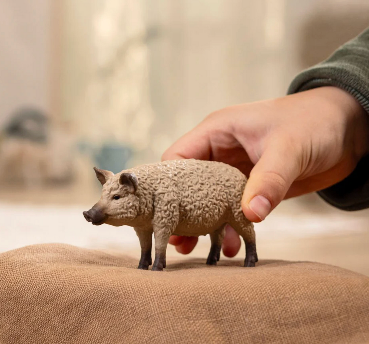Hand holding a small, curly-haired Mangalica Pig figurine with a textured gray back on a brown surface.