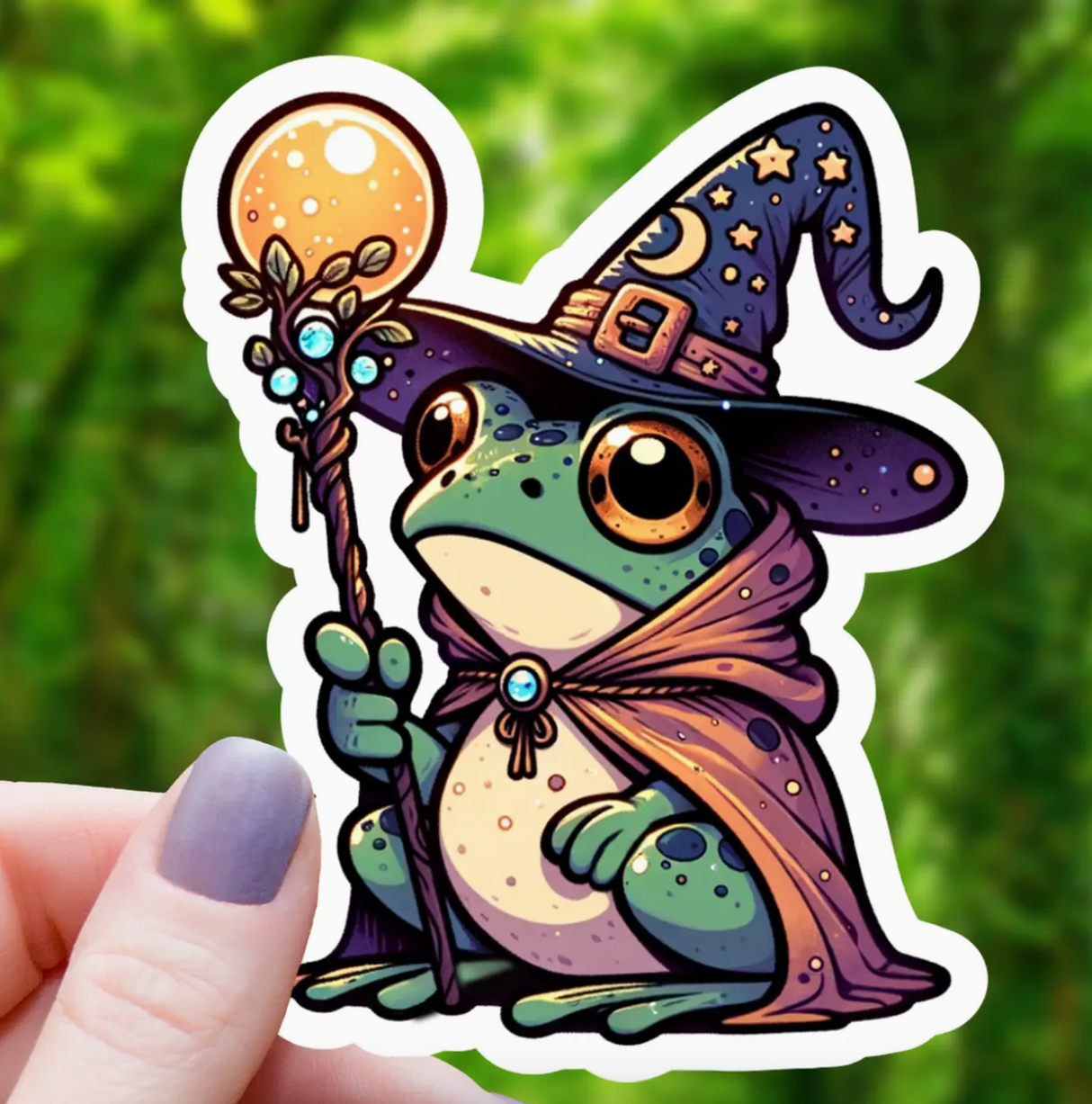 Hand holding a colorful sticker of a frog dressed as a wizard with a hat, cloak, and magical staff outdoors.