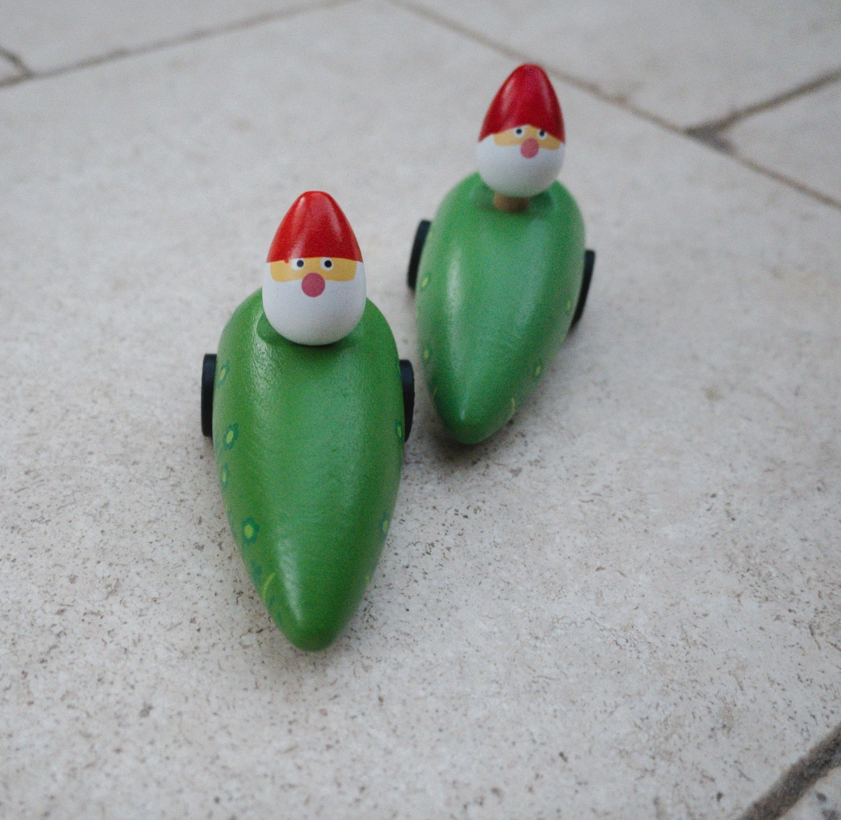 Two wooden garden gnomes with red hats seated in green pullback cars on a tiled floor.