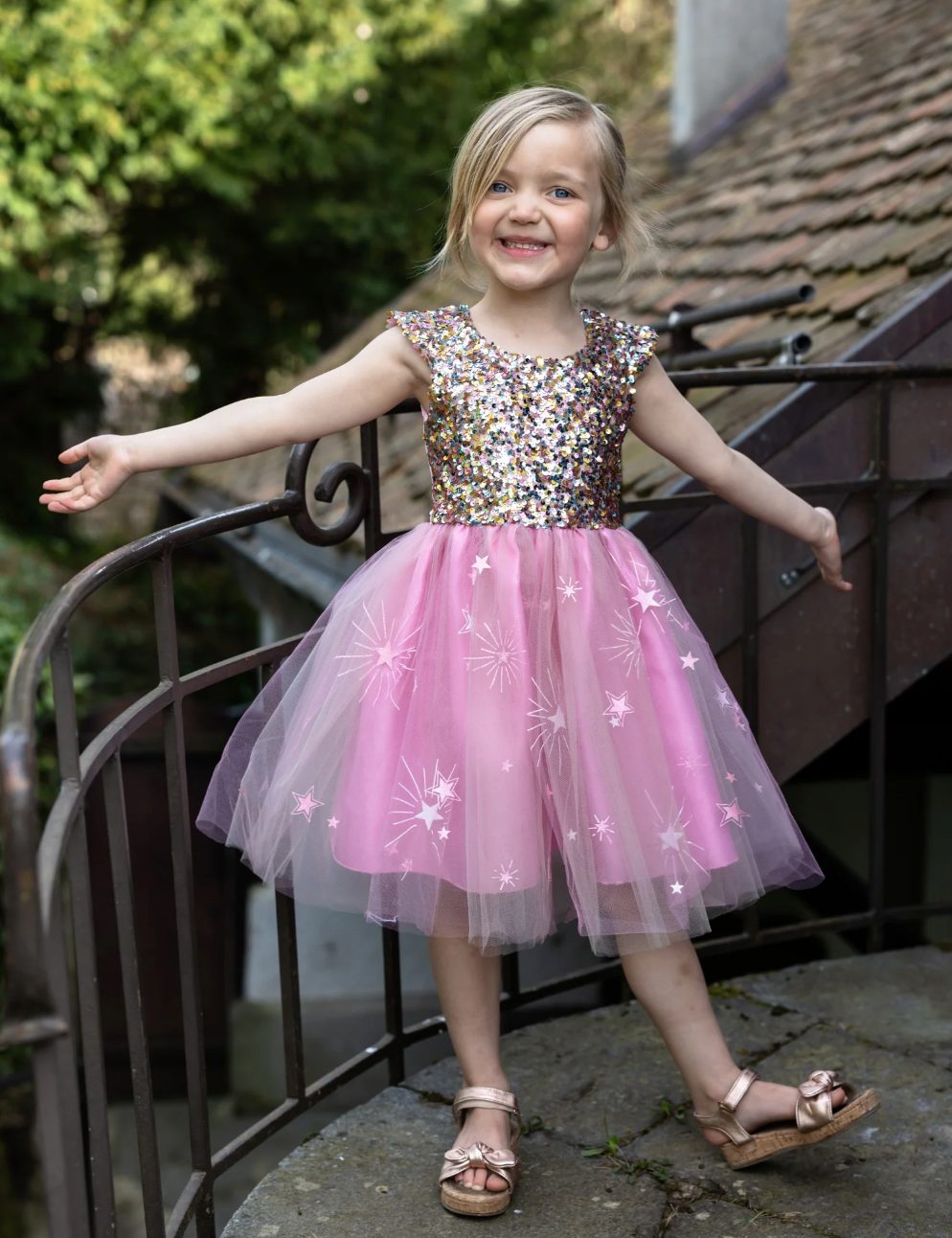 Girl wearing a multicolored sequin bodice dress with a pink tulle skirt decorated with stars, smiling outdoors on stairs