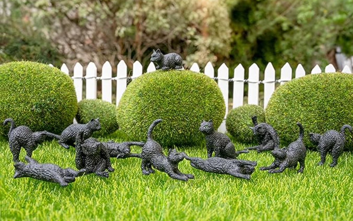 Twelve miniature black cat figurines in various playful poses arranged on grass with a white picket fence background