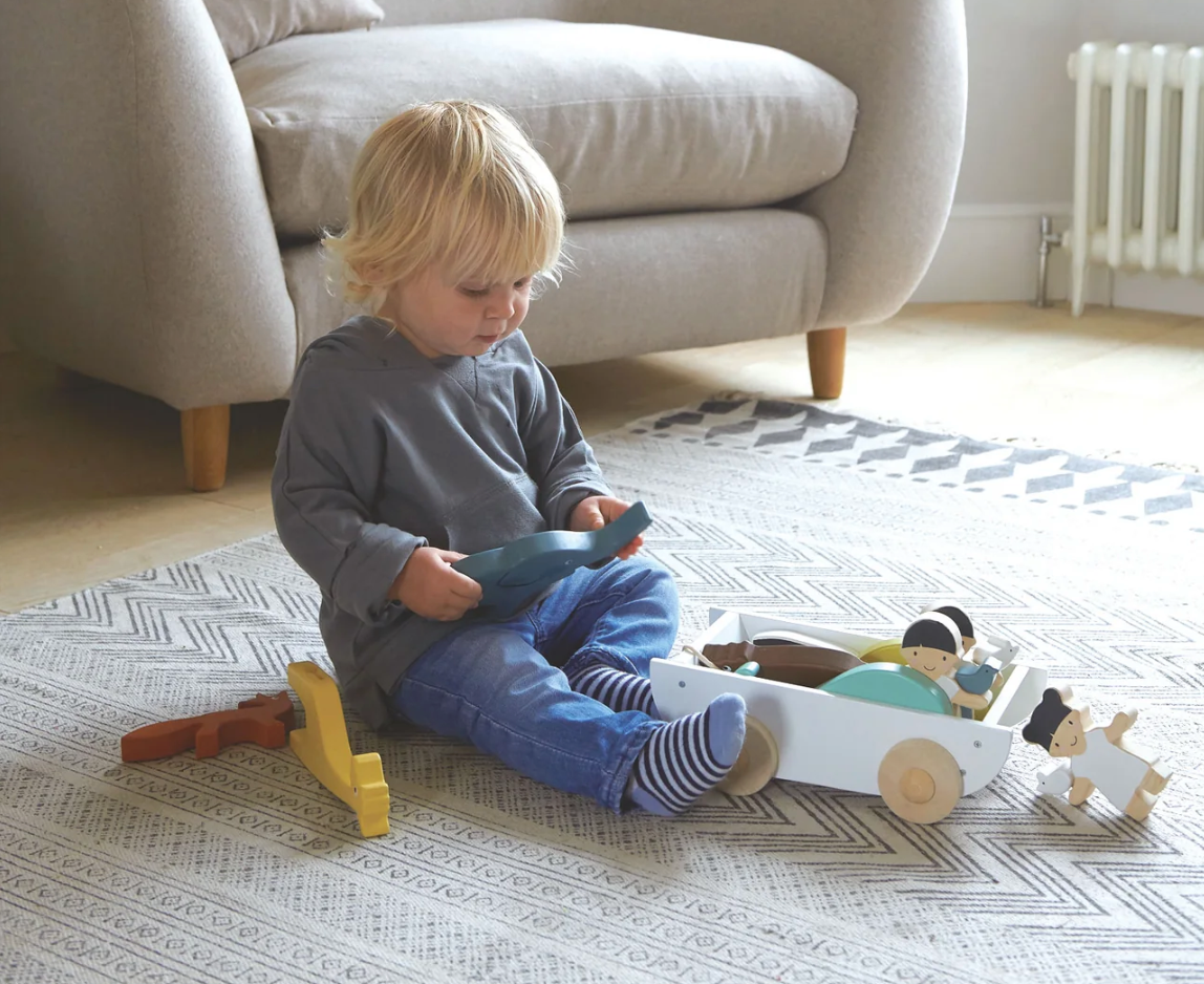 Toddler playing on a rug with a white wooden pull toy boat and colorful animal figures including a boy and girl.
