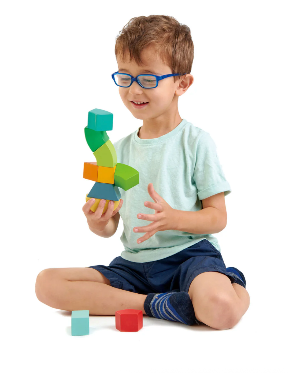 Young boy wearing blue glasses playing with colorful magnetic wooden blocks while sitting cross-legged on floor