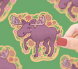 Hand holding a cute purple moose sticker surrounded by autumn leaves, pinecones, and flowers on green background