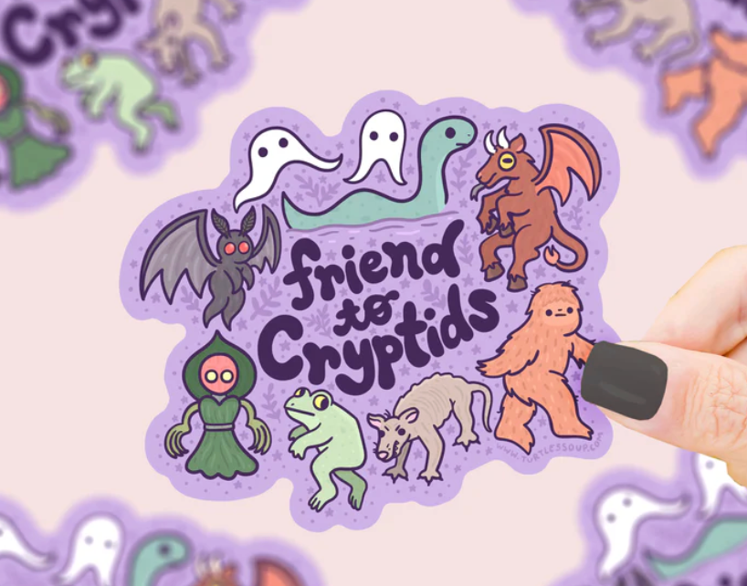 Hand holding a colorful "Friend to Cryptids" sticker featuring friendly cartoon cryptids on a purple background