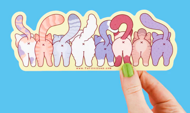 Hand holding a colorful sticker showing the backsides of seven different cartoon cats with raised tails on blue background
