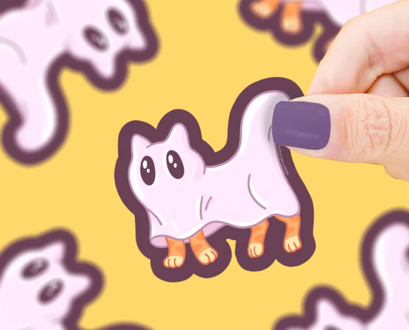 Hand holding a cute ghost kitty sticker with purple nails on a yellow background with more stickers.