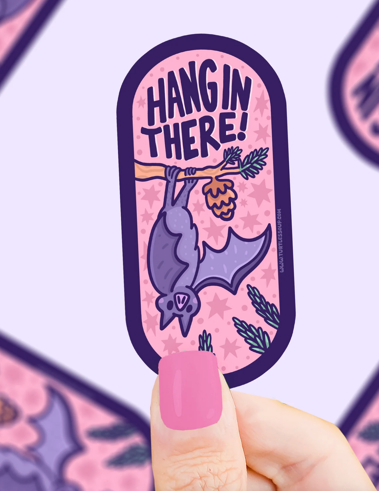 Hand holding a purple bat sticker hanging upside down from a branch with the words Hang in There on pink background