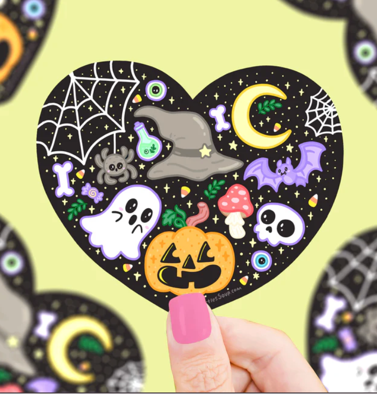 Hand holding a heart-shaped Halloween sticker featuring ghost, pumpkin, bat, spider, moon, and potion bottle designs.
