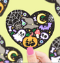 Hand holding a heart-shaped Halloween sticker featuring ghost, pumpkin, bat, spider, moon, and potion bottle designs.