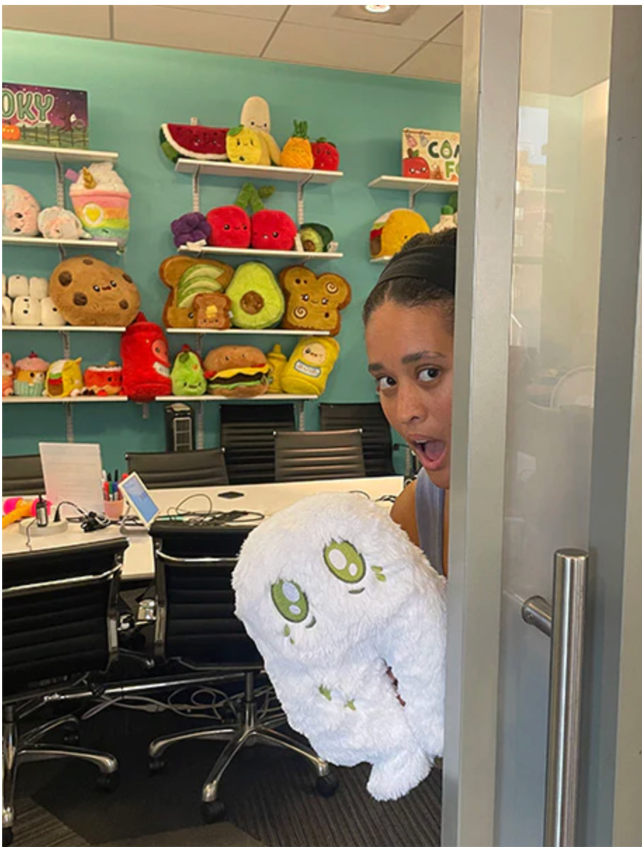 Person peeks from behind a door holding a fluffy white plush toy with green eyes in a colorful office filled with stuffed toys.