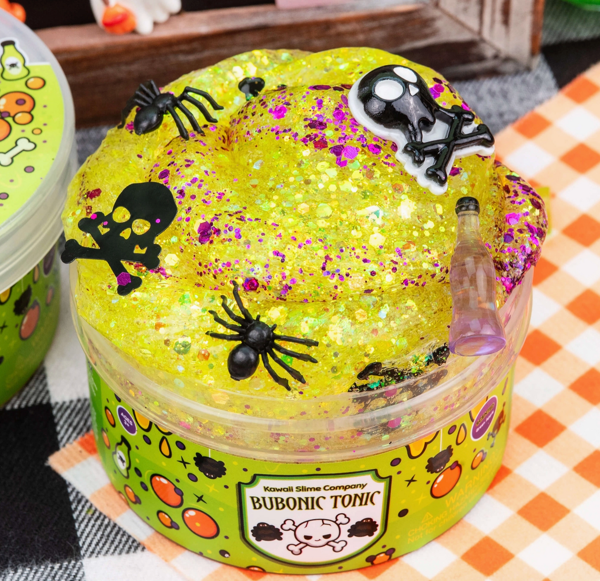 Clear yellow glitter slime with spooky skulls, black spiders, and a small potion bottle on top in a labeled container