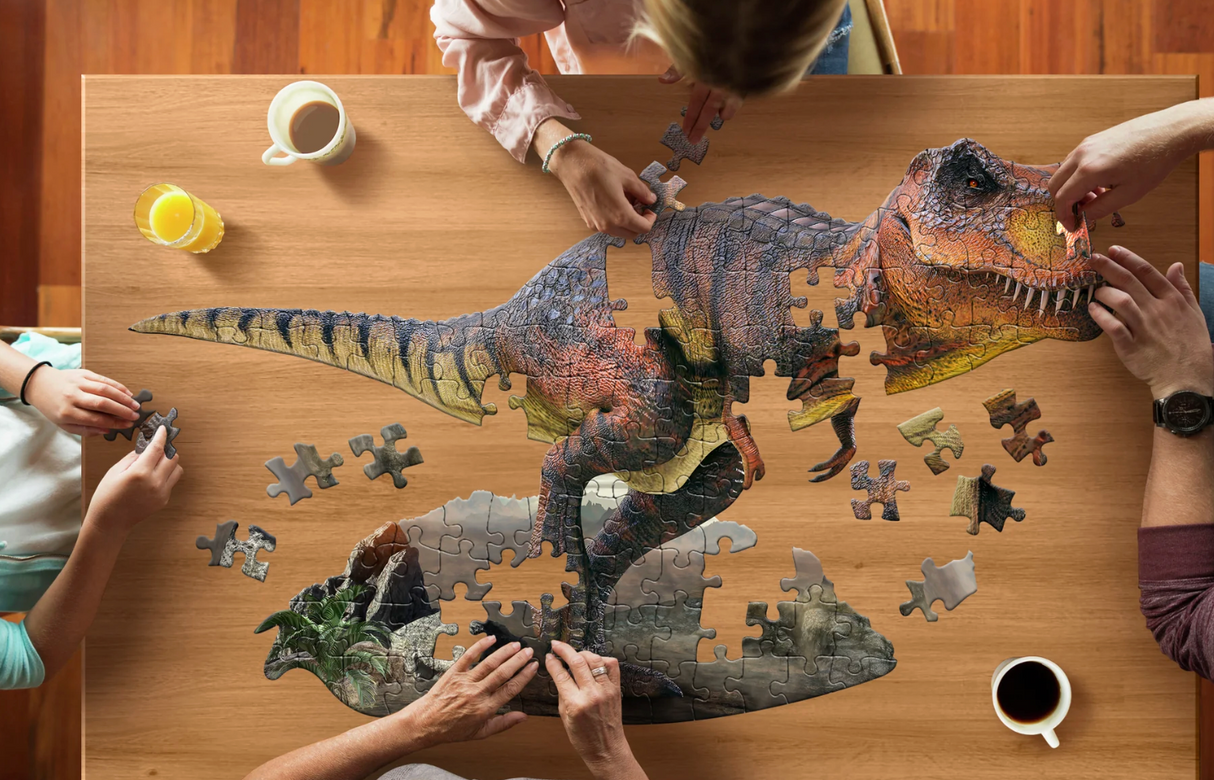 People assembling a large 100-piece dinosaur-shaped jigsaw puzzle on a wooden table with drinks nearby