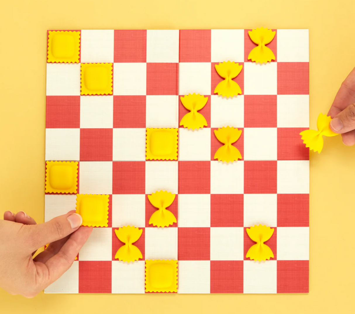 Hands playing a checkers game with pasta-shaped pieces on a red-and-white checkered board