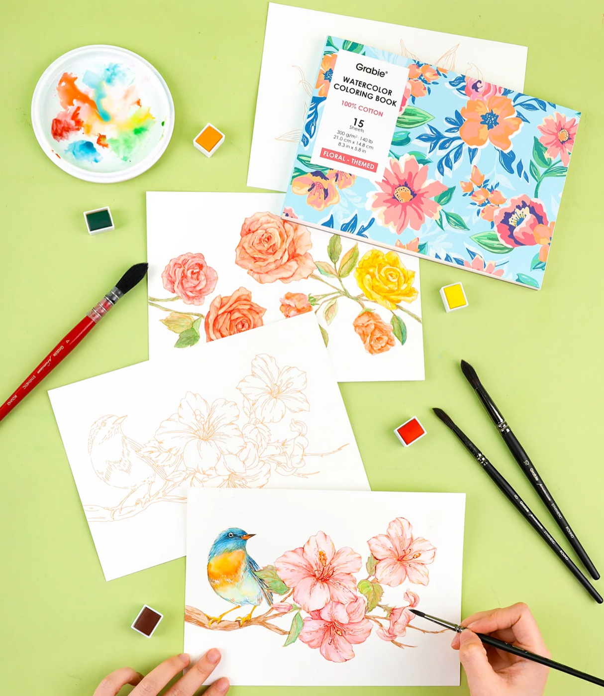 Watercolor coloring book with floral designs, paintbrushes, and small watercolor palettes on a green background
