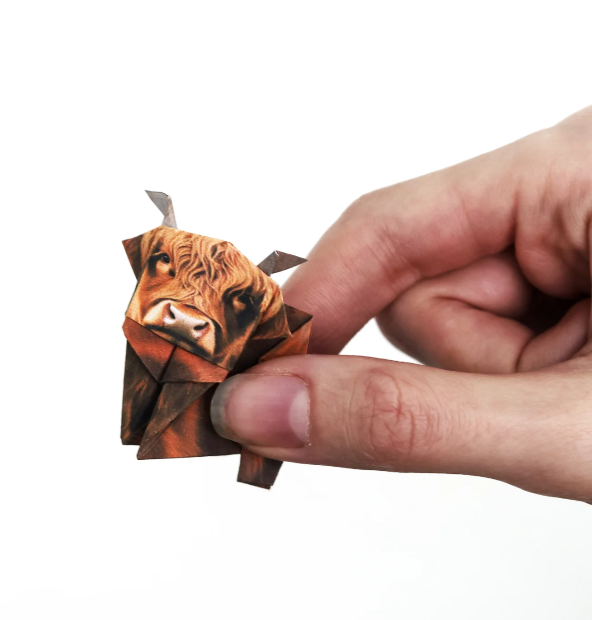 Hand holding a small origami Highland cow made from a pre-printed paper folding kit with detailed face design