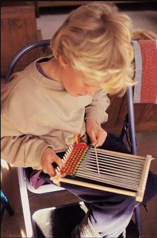 Young person weaving colorful yarn on a hardwood peg loom with cotton warp strings in a cozy indoor setting