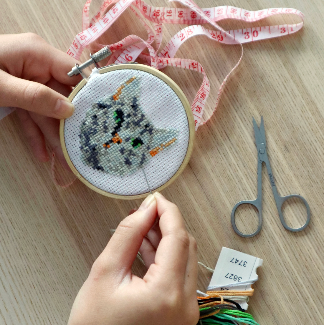 Hands stitching a mini embroidery hoop with a cute gray cat design on white fabric on a wooden table