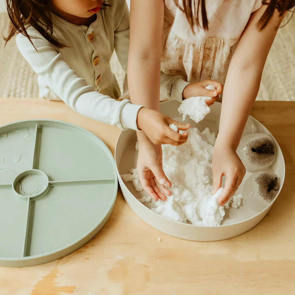 Two children playing with white sensory snow in a round container on a wooden table indoors